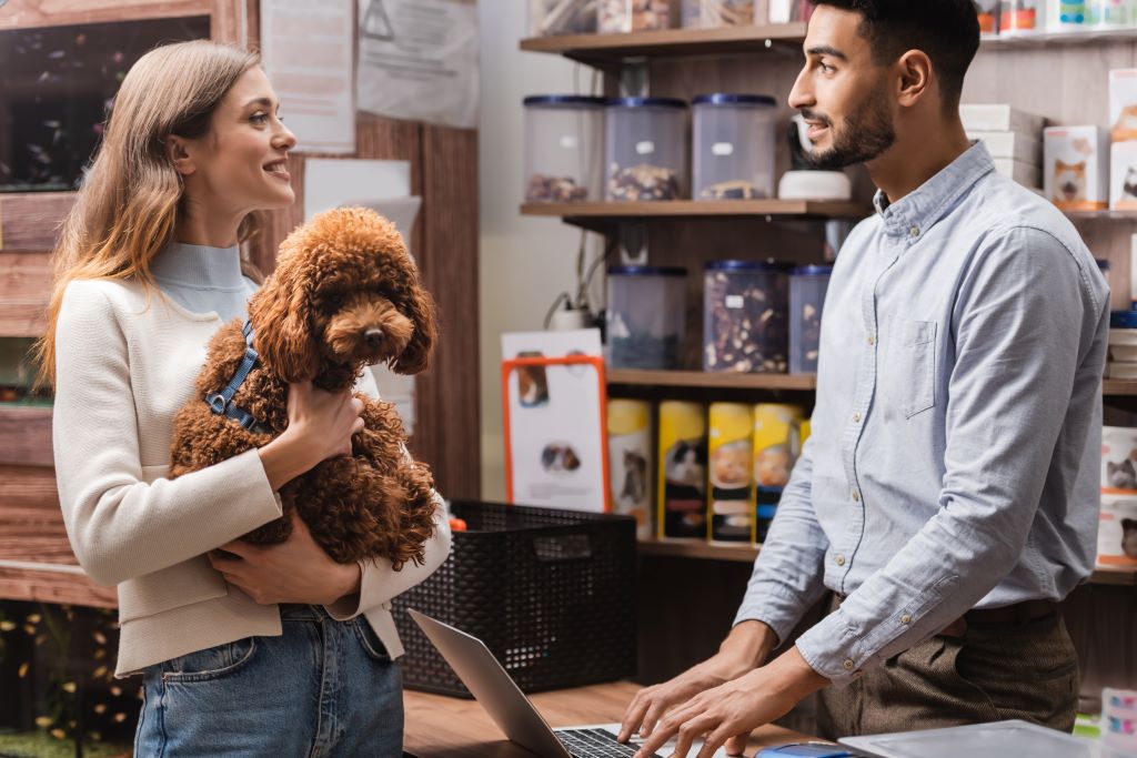 Na foto há uma mulher loira segurando um cachorro peludo marrom no colo, enquanto conversa com um homem que está atrás do balcão, na frente de um notebook, ambos estão conversando e sorrindo, ao que parece a mulher está finalizando alguma compra em uma distribuidora pet.