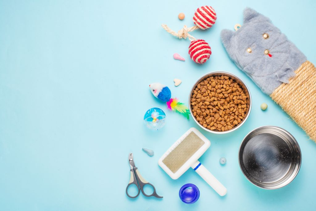 Na imagem, um fundo azul claro e na frente, uma série de produtos de pet shop, tais coo escova para animais, brinquedos, um pote cheio de ração, bolinhas e um cortador de unha pet.