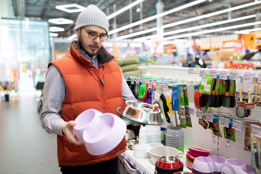 Na foto, um homem vestido com um colete laranjado e touca está segurando dois comedouros de cachorro e olhando pra eles em dúvida de qual escolher. O homem está em uma distribuidora de produtos para pet.