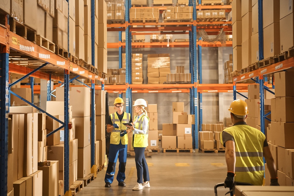 A imagem é de um corredor de uma distribuidora, neste corredor estão 3 fujncionários, ambos com capacete e colete.